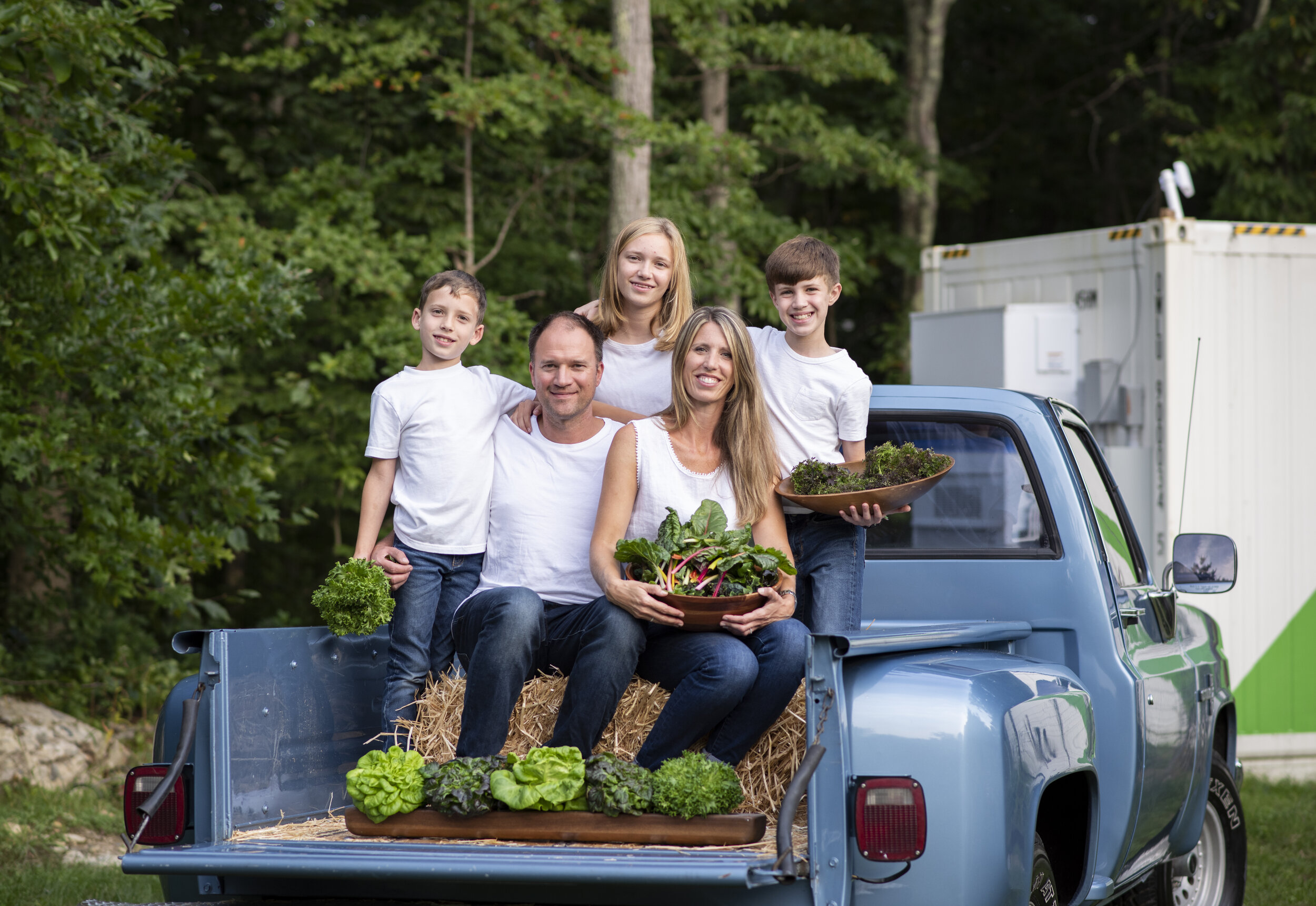 Family enjoying fresh vegetables in a pickup truck, surrounded by greenery. Parents and three children hold bowls of harvested produce, showcasing a healthy lifestyle and home gardening.